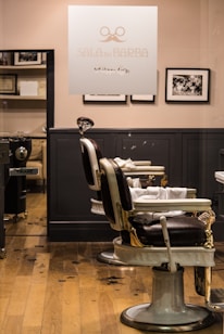 A barbershop interior features vintage-style barber chairs with white and black leather, positioned in front of a wooden floor. The walls are adorned with black-and-white photographs, and a glass panel with the logo and name 'SALA DE BARBA' provides a view into the well-organized space.