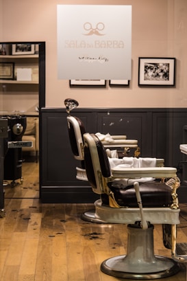 A barbershop interior features vintage-style barber chairs with white and black leather, positioned in front of a wooden floor. The walls are adorned with black-and-white photographs, and a glass panel with the logo and name 'SALA DE BARBA' provides a view into the well-organized space.