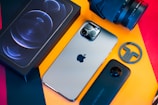 A flat-lay image of a smartphone with a sleek design, a camera lens, a smartphone box, a circular logo piece, and what appears to be a camera case all arranged on a vibrant background with orange and red colors.