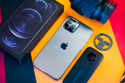 A flat-lay image of a smartphone with a sleek design, a camera lens, a smartphone box, a circular logo piece, and what appears to be a camera case all arranged on a vibrant background with orange and red colors.