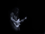 A person plays an electric guitar with a focused expression, set against a dark, minimalist background. The guitar is illuminated, highlighting its white or light-colored body and strings.
