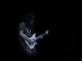 A person plays an electric guitar with a focused expression, set against a dark, minimalist background. The guitar is illuminated, highlighting its white or light-colored body and strings.