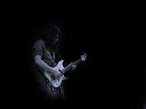 A person plays an electric guitar with a focused expression, set against a dark, minimalist background. The guitar is illuminated, highlighting its white or light-colored body and strings.