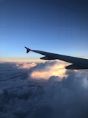 Commercial airplane wing soaring above fluffy white clouds during sunrise.