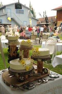 An outdoor gathering features a rustic cake display with three white frosted cakes adorned with yellow flowers on wooden stands. In the background, people socialize near a blue building with balloons, and photographs are hung on a line.