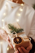 A person holds a beautifully wrapped gift adorned with natural elements such as a sprig of evergreen, a dried orange slice, and string. The package is wrapped in plain brown paper and decorated with sheet music, set against a softly lit background with warm, glowing lights.