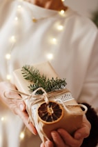 A person holds a beautifully wrapped gift adorned with natural elements such as a sprig of evergreen, a dried orange slice, and string. The package is wrapped in plain brown paper and decorated with sheet music, set against a softly lit background with warm, glowing lights.