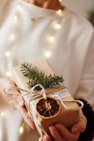 A person holds a beautifully wrapped gift adorned with natural elements such as a sprig of evergreen, a dried orange slice, and string. The package is wrapped in plain brown paper and decorated with sheet music, set against a softly lit background with warm, glowing lights.
