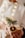 A person holds a beautifully wrapped gift adorned with natural elements such as a sprig of evergreen, a dried orange slice, and string. The package is wrapped in plain brown paper and decorated with sheet music, set against a softly lit background with warm, glowing lights.