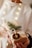 A person holds a beautifully wrapped gift adorned with natural elements such as a sprig of evergreen, a dried orange slice, and string. The package is wrapped in plain brown paper and decorated with sheet music, set against a softly lit background with warm, glowing lights.