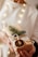 A person holds a beautifully wrapped gift adorned with natural elements such as a sprig of evergreen, a dried orange slice, and string. The package is wrapped in plain brown paper and decorated with sheet music, set against a softly lit background with warm, glowing lights.