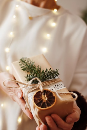 A person holds a beautifully wrapped gift adorned with natural elements such as a sprig of evergreen, a dried orange slice, and string. The package is wrapped in plain brown paper and decorated with sheet music, set against a softly lit background with warm, glowing lights.