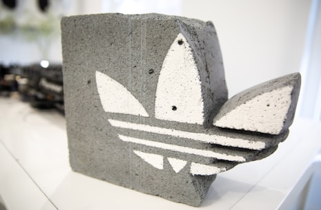 A concrete sculpture shaped like a stylized leaf pattern with three distinct white leaves set against a gray background. It sits on a smooth, white surface with a blurred background, hinting at an indoor setting.