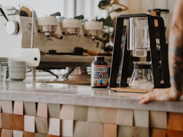 A coffee shop countertop features a modern coffee brewing setup and a container labeled 'Redhats Colombia'. In the background, a large espresso machine is visible next to a stack of saucers. The setting is warm and inviting, with a hand resting on the counter to the right.
