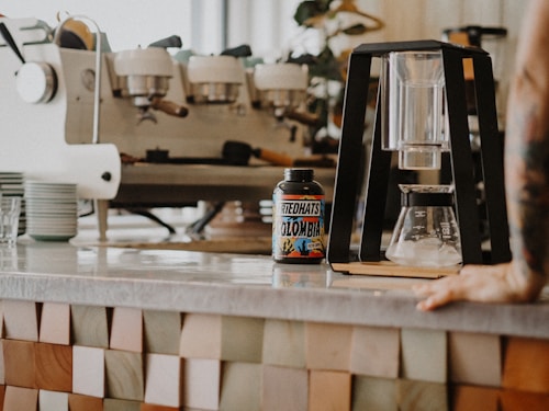 A coffee shop countertop features a modern coffee brewing setup and a container labeled 'Redhats Colombia'. In the background, a large espresso machine is visible next to a stack of saucers. The setting is warm and inviting, with a hand resting on the counter to the right.