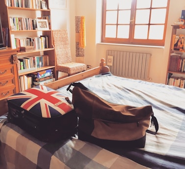 A cozy bedroom features a bed with a checkered bedspread, a suitcase with a Union Jack design, and a brown leather bag on top. Bookshelves filled with books line the wall, and a wicker chair sits beside them. Sunlight streams in through a large window, illuminating the room.