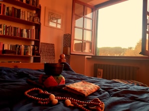 A cozy bedroom scene with warm lighting, featuring a neatly arranged bed with a blue blanket. A singing bowl sits on a cushion, accompanied by a beaded necklace and folded fabric. A wall-mounted bookshelf filled with books adorns the left side, while an open window on the right offers a view of trees during sunset.