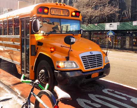 A bright yellow school bus is parked on a city street. The bus has noticeable black detailing and is equipped with safety mirrors. In front of the bus, a bicycle with a frame and wheel partly in view is parked. The setting appears urban, with buildings in the background and a storefront partially visible.