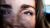 A close-up of a person's eyes and eyebrows, with strands of hair partially covering the face. The focus is on the person's expressive gaze, with natural skin texture visible.