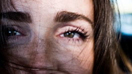 A close-up of a person's eyes and eyebrows, with strands of hair partially covering the face. The focus is on the person's expressive gaze, with natural skin texture visible.