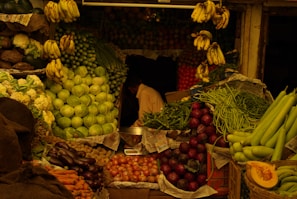 A vibrant fruit and vegetable market stall displaying a variety of fresh produce. There are piles of cabbages, bananas hanging in clusters, cauliflowers, eggplants, carrots, tomatoes, long beans, and other vegetables. The stall is densely packed and organized in an inviting manner, with newsprint used to line the produce. A person is partially visible behind the stall, engaged in sorting or arranging the items.