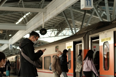 Passengers are standing on a train platform, some boarding and others waiting while using their mobile phones. The platform has a modern architectural design with metallic beams and a partially transparent roof. People are wearing masks, indicating an awareness of health safety.