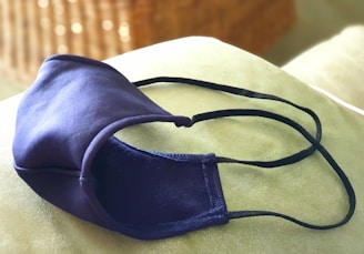 A dark blue cloth face mask rests on a light green surface. The mask has black ear straps and its texture appears soft. The background is blurred, featuring soft brown and tan tones that suggest a wicker or woven surface.