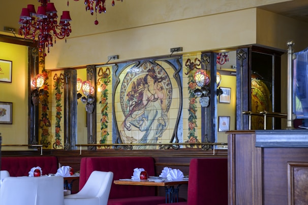 An elegant interior space showcasing a large decorative mural with an Art Nouveau style depicting a woman surrounded by floral patterns. Vibrant red chandeliers hang from the ceiling, while the seating area features plush red and white chairs and tables set with red candles and neatly folded napkins.
