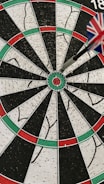 A dartboard with a dart accurately hitting the bullseye. The dart's flight has a pattern resembling the Union Jack. The board is segmented with alternating colors of red, green, black, and white.