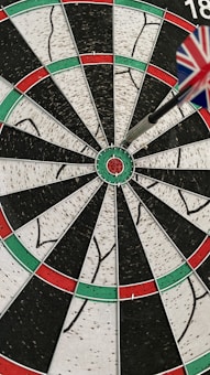 A dartboard with a dart accurately hitting the bullseye. The dart's flight has a pattern resembling the Union Jack. The board is segmented with alternating colors of red, green, black, and white.
