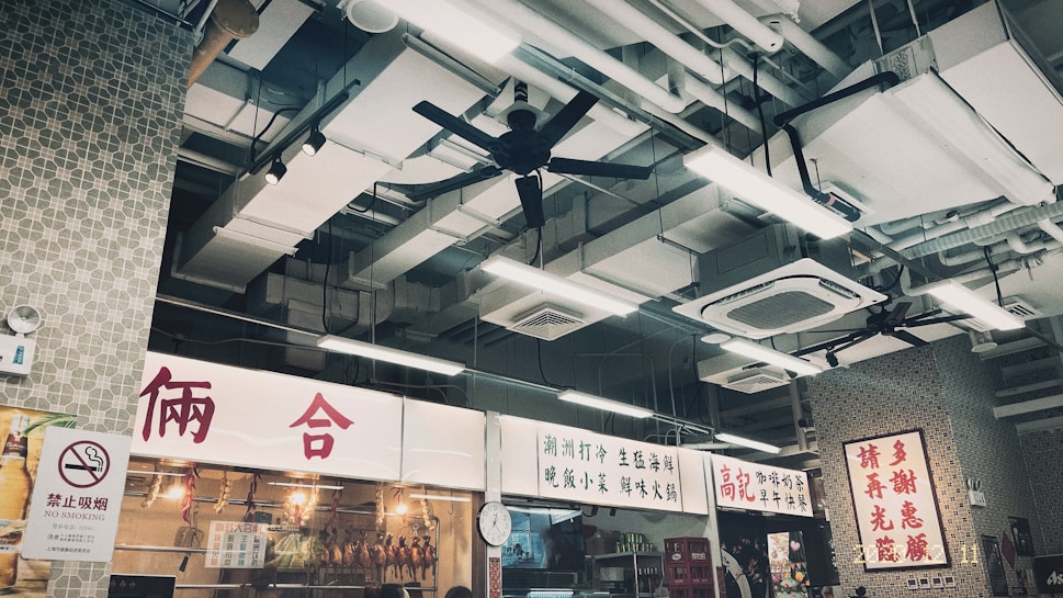 An indoor market setting featuring several overhead ceiling fans and industrial lighting. Signs with Chinese characters are prominently displayed, along with a 'No Smoking' sign. The ceiling exhibits a complex structure of pipes and air conditioning units.