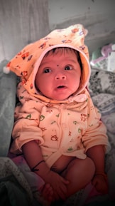 A baby dressed in a cozy outfit with a hood decorated with small animal prints. The baby is looking upwards with wide eyes, sitting on a patterned fabric. The hood features a bear design, and the baby has a small black dot on the forehead. The background includes some soft items, possibly a blanket or pillow.