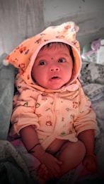A baby dressed in a cozy outfit with a hood decorated with small animal prints. The baby is looking upwards with wide eyes, sitting on a patterned fabric. The hood features a bear design, and the baby has a small black dot on the forehead. The background includes some soft items, possibly a blanket or pillow.