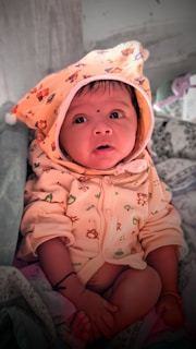 A baby dressed in a cozy outfit with a hood decorated with small animal prints. The baby is looking upwards with wide eyes, sitting on a patterned fabric. The hood features a bear design, and the baby has a small black dot on the forehead. The background includes some soft items, possibly a blanket or pillow.