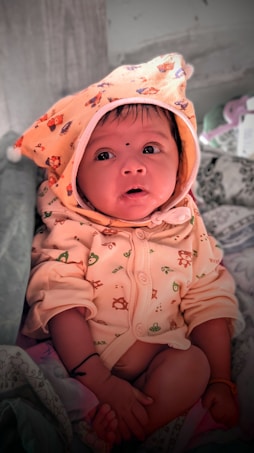 A baby dressed in a cozy outfit with a hood decorated with small animal prints. The baby is looking upwards with wide eyes, sitting on a patterned fabric. The hood features a bear design, and the baby has a small black dot on the forehead. The background includes some soft items, possibly a blanket or pillow.