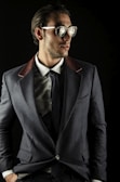 A man wearing sunglasses, a gray suit jacket with contrasting lapels, a white dress shirt, and a dark tie. The setting is against a dark background, giving a fashionable and sophisticated appearance.