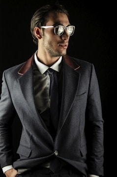 A man wearing sunglasses, a gray suit jacket with contrasting lapels, a white dress shirt, and a dark tie. The setting is against a dark background, giving a fashionable and sophisticated appearance.