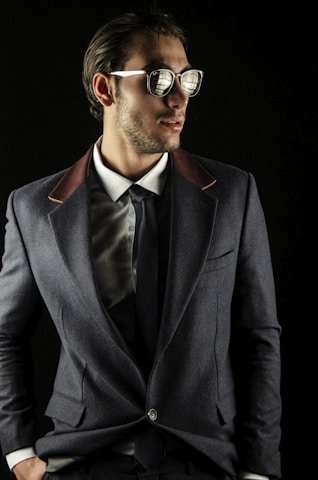 A man wearing sunglasses, a gray suit jacket with contrasting lapels, a white dress shirt, and a dark tie. The setting is against a dark background, giving a fashionable and sophisticated appearance.