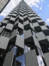 A striking architectural structure featuring a series of patterned, textured panels on its facade, extending upwards into the sky with a dynamic and geometric design. The building showcases a modern, innovative look with reflective glass segments interspersed between the panels.