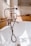 A bathroom scene featuring a vintage-style bronze bathtub faucet with a handheld shower attachment and flexible hose, set against a white bathtub. The background includes a wooden surface with decorative items like a round dish and textured white vase. Natural light streams in from a nearby window, casting soft shadows.
