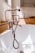 A bathroom scene featuring a vintage-style bronze bathtub faucet with a handheld shower attachment and flexible hose, set against a white bathtub. The background includes a wooden surface with decorative items like a round dish and textured white vase. Natural light streams in from a nearby window, casting soft shadows.