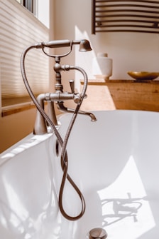 A bathroom scene featuring a vintage-style bronze bathtub faucet with a handheld shower attachment and flexible hose, set against a white bathtub. The background includes a wooden surface with decorative items like a round dish and textured white vase. Natural light streams in from a nearby window, casting soft shadows.