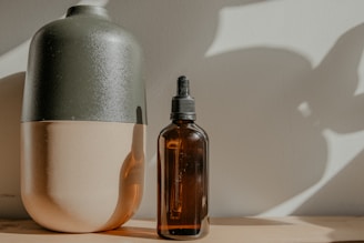 A dark green and beige ceramic vase is placed next to a brown glass bottle with a dropper. Both objects are resting on a wooden surface with soft, natural light casting shadows on the wall behind them.