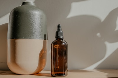 A dark green and beige ceramic vase is placed next to a brown glass bottle with a dropper. Both objects are resting on a wooden surface with soft, natural light casting shadows on the wall behind them.