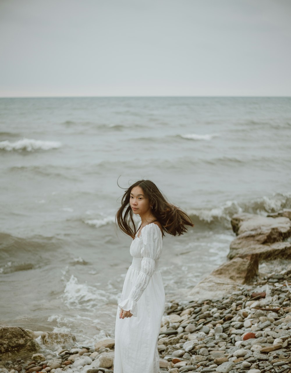 woman in white long sleeve dress standing on brown rock near sea during daytime