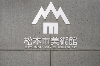 A grey concrete wall features a minimalist, geometric logo resembling the letter 'M' positioned above Japanese characters and English text that reads 'Matsumoto City Museum of Art'.