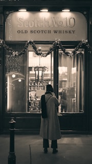 A person stands in front of a whiskey shop window adorned with festive garlands. The sign above the window reads 'Scotch No10 Old Scotch Whisky.' The scene has a vintage or classic atmosphere.