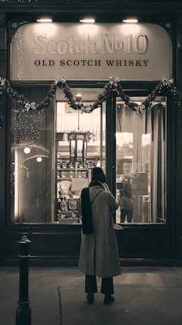 A person stands in front of a whiskey shop window adorned with festive garlands. The sign above the window reads 'Scotch No10 Old Scotch Whisky.' The scene has a vintage or classic atmosphere.