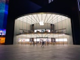 A modern, large glass-fronted store with an illuminated Apple logo on the facade. The interior features two levels visible through the clear glass with various Apple products on display. People are seen inside and outside the store, some walking and others standing.