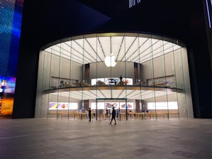 A modern, large glass-fronted store with an illuminated Apple logo on the facade. The interior features two levels visible through the clear glass with various Apple products on display. People are seen inside and outside the store, some walking and others standing.